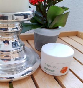 CBD Akutbalsam von CBD VITAL im Test 2020 | Biokosmetik mit CBD-Superkraft 1 Eine Dose CBD-Akutbalsam von CBDvital steht auf einen Holztisch neben einen Kerzenständer und einer roten Topfplanze